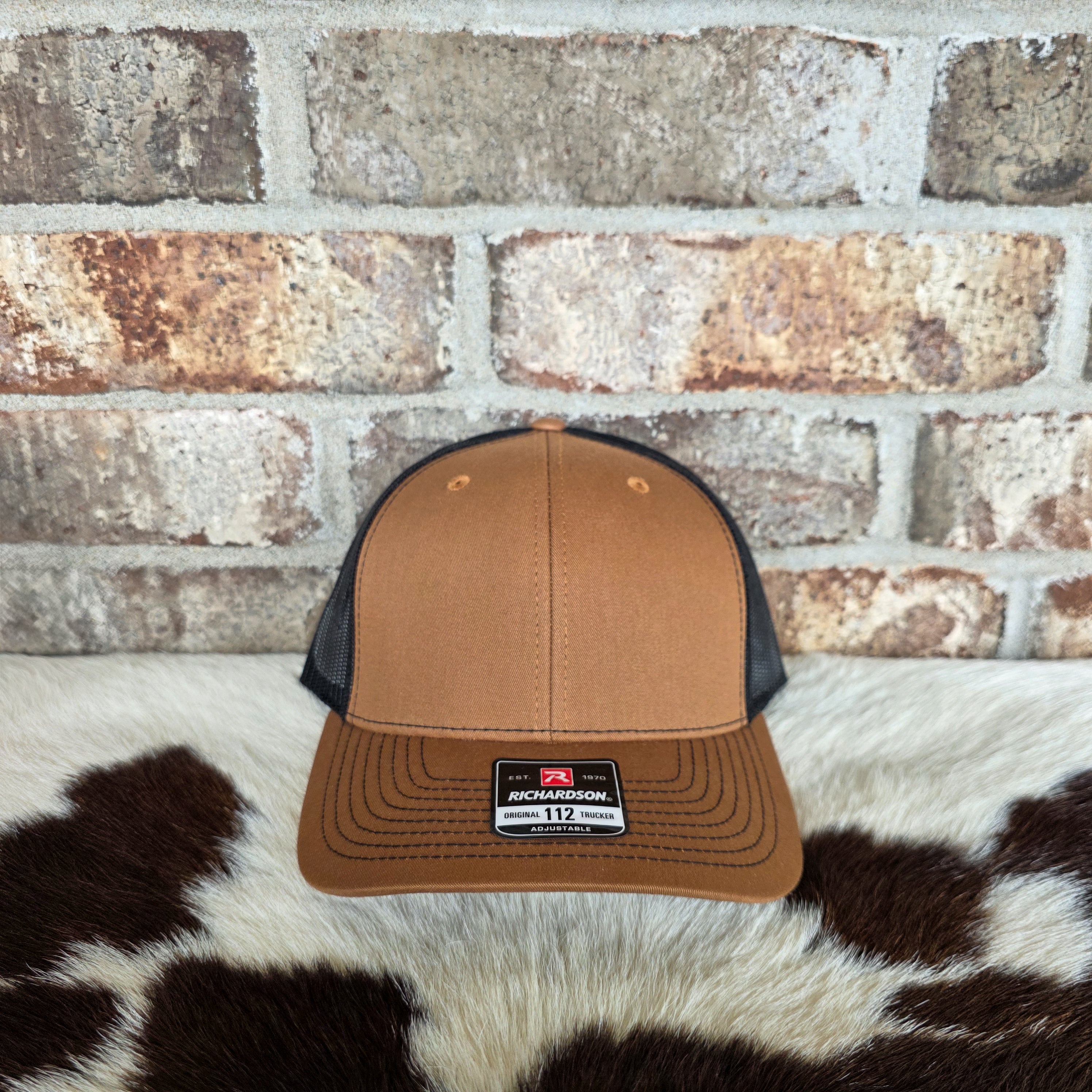 Brown and black trucker hat with Richardson logo on a cowhide rug against a brick wall.