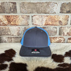 Blue and gray cap with a brand label on a cowhide rug against a brick wall.