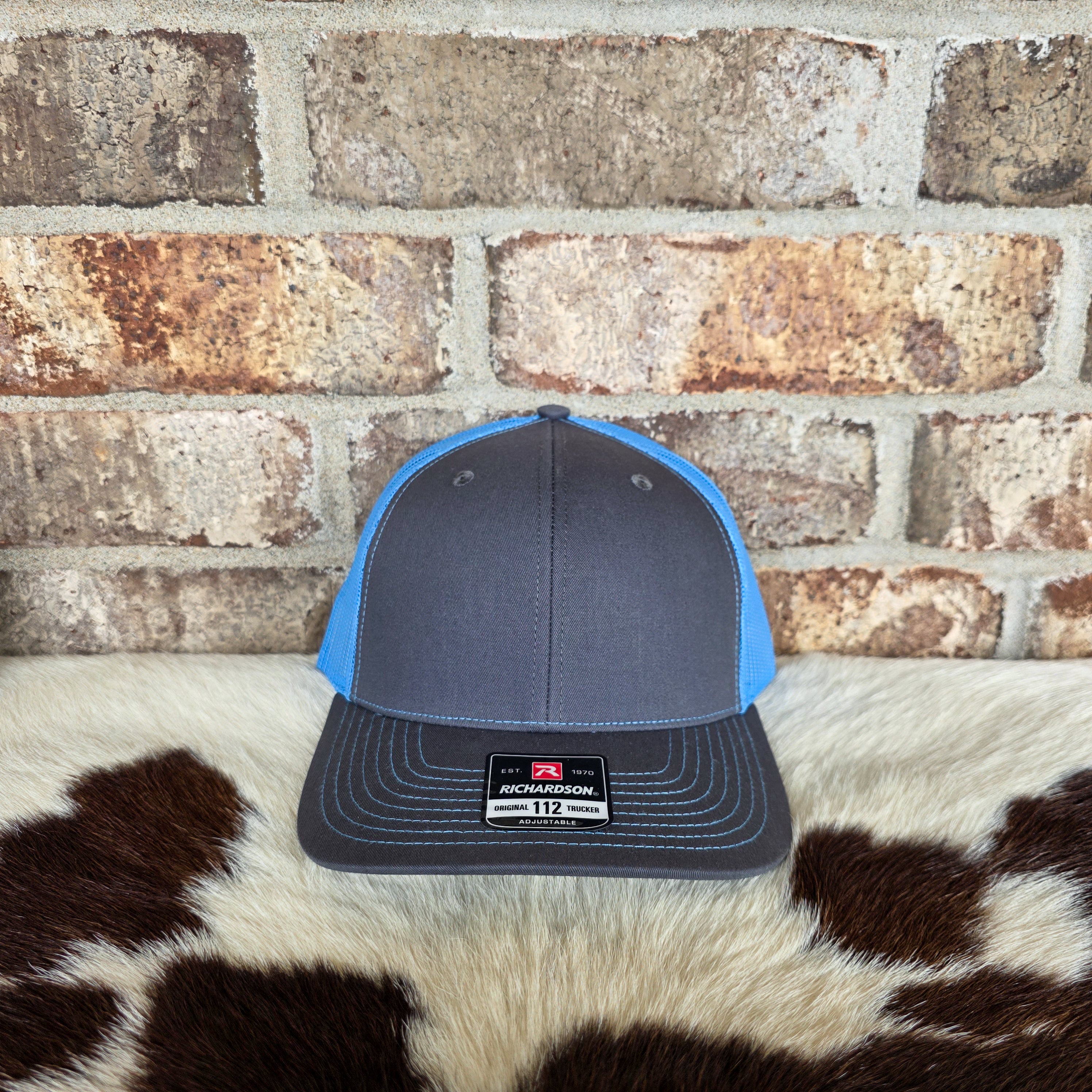 Blue and gray cap with a brand label on a cowhide rug against a brick wall.