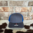 Gray and blue cap with a Richardson logo on a cowhide rug against a brick wall.