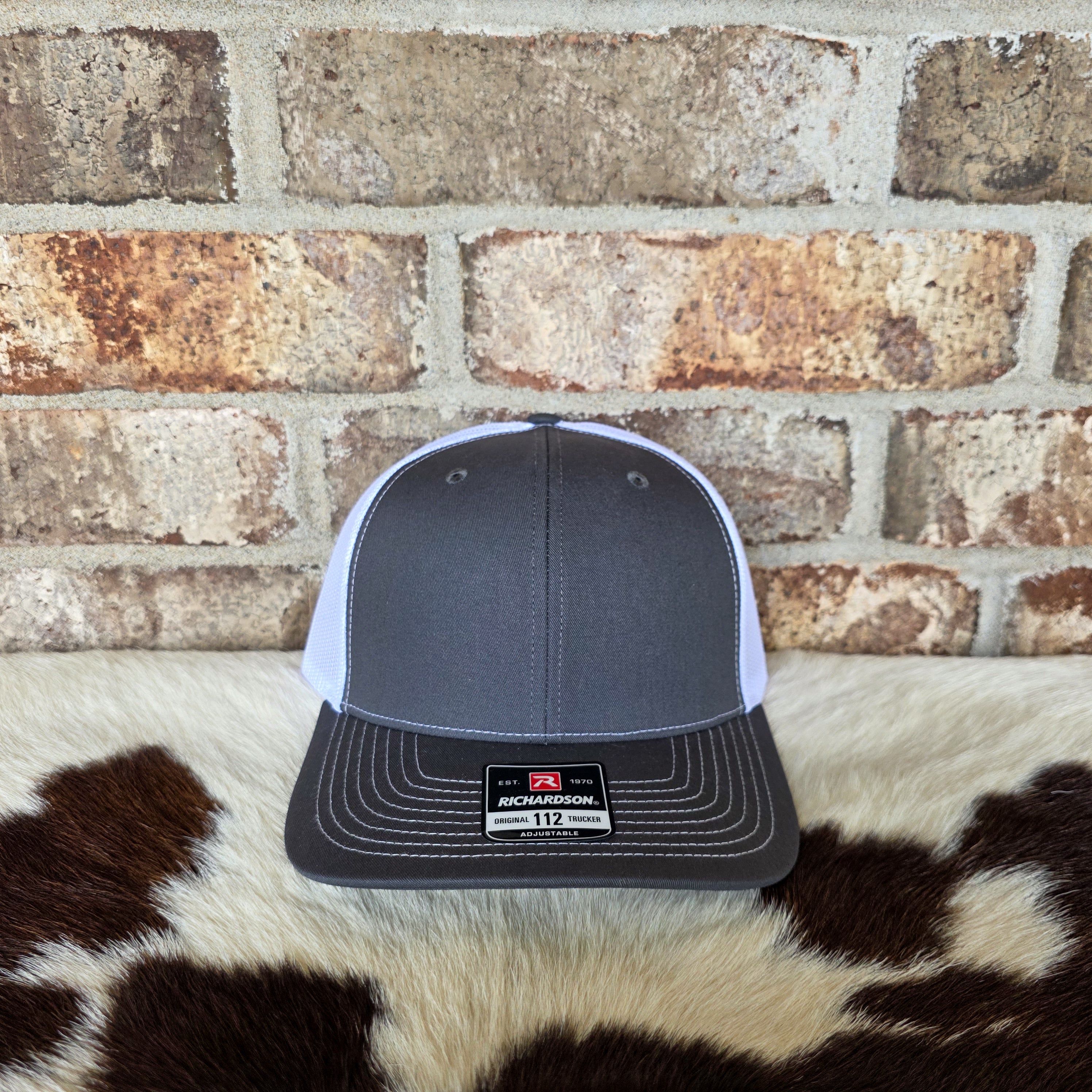 Gray and white cap with a Richardson logo on a cowhide rug against a brick wall.