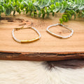 One gold bar and one silver bar bracelet with opalescent white beads are displayed on a rustic wooden slice, with green eucalyptus leaves and cowhide in the background.