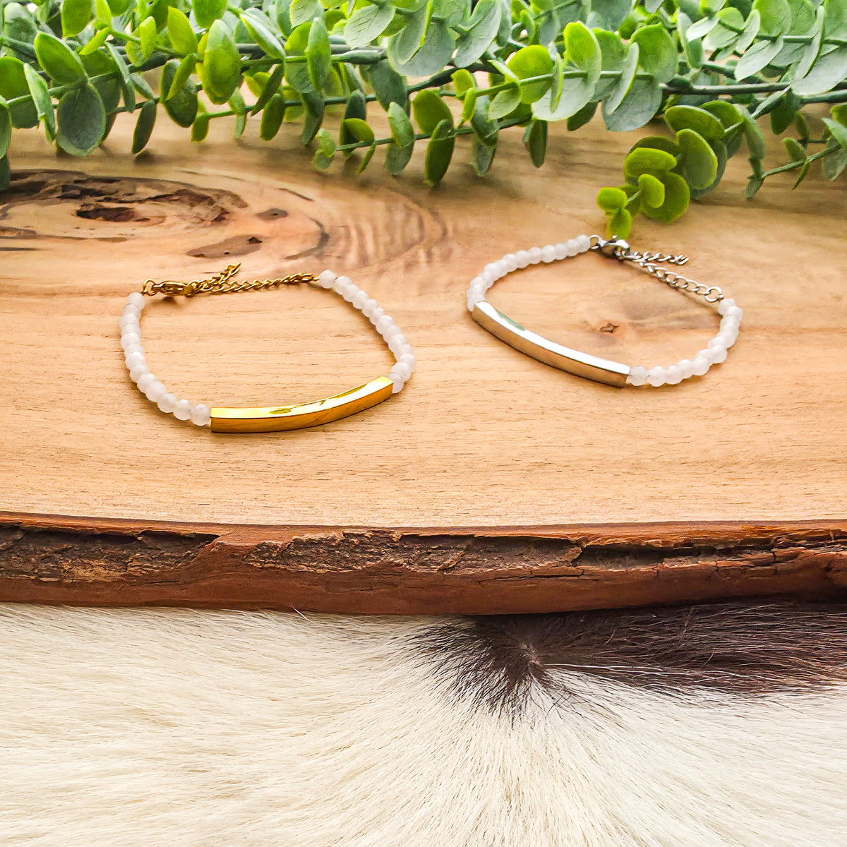 One gold bar and one silver bar bracelet with opalescent white beads are displayed on a rustic wooden slice, with green eucalyptus leaves and cowhide in the background.