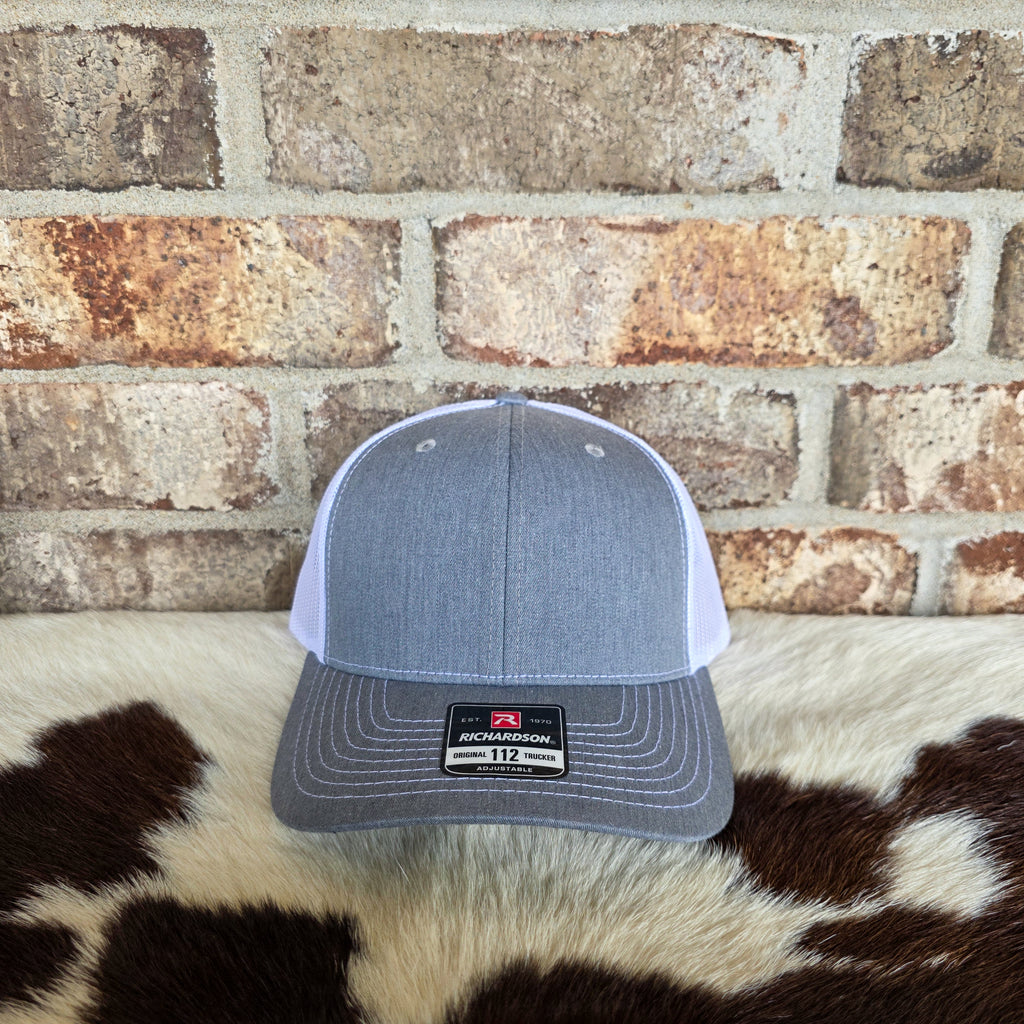 Gray and white cap with Richardson logo on a cowhide rug against a brick wall.
