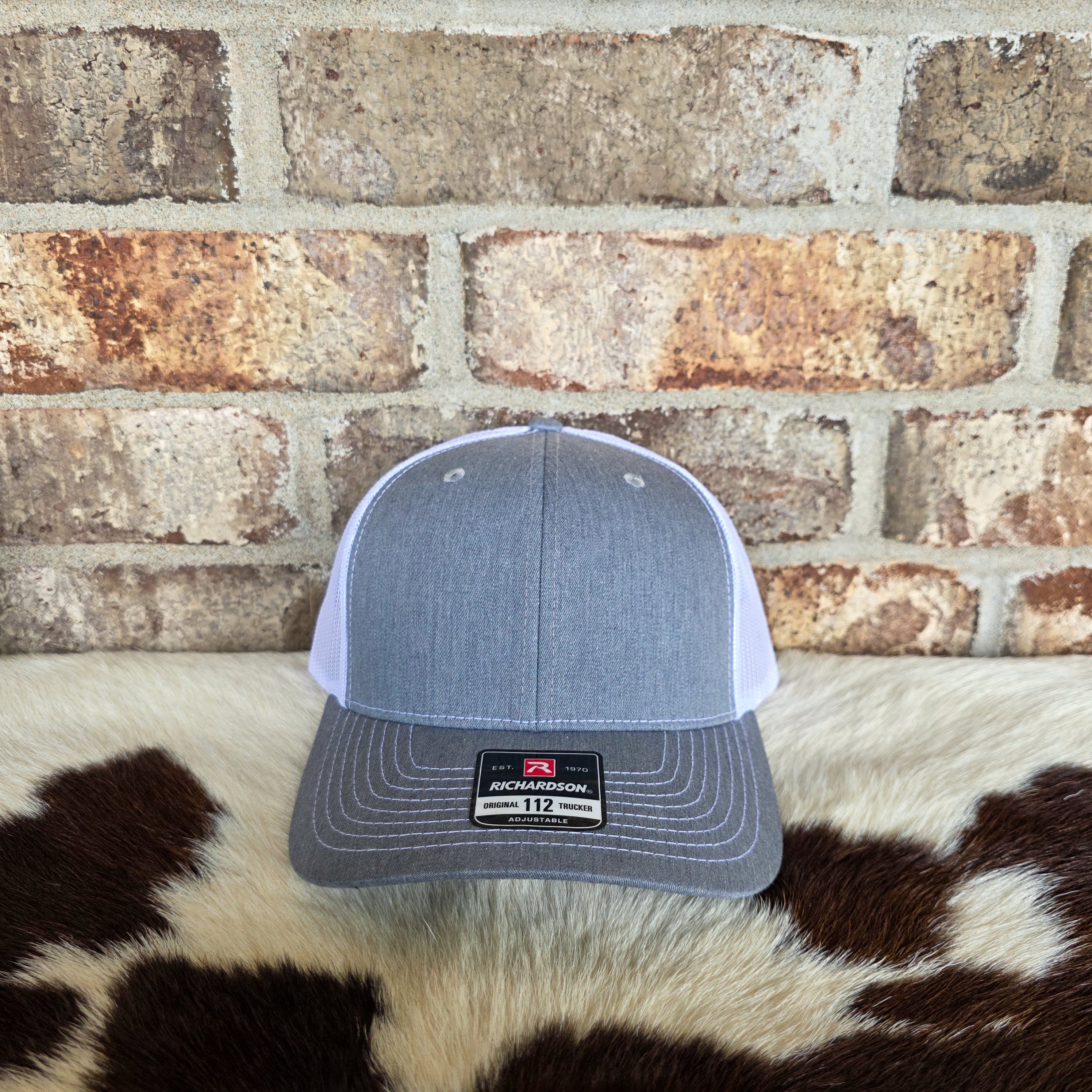 Gray and white cap with Richardson logo on a cowhide rug against a brick wall.