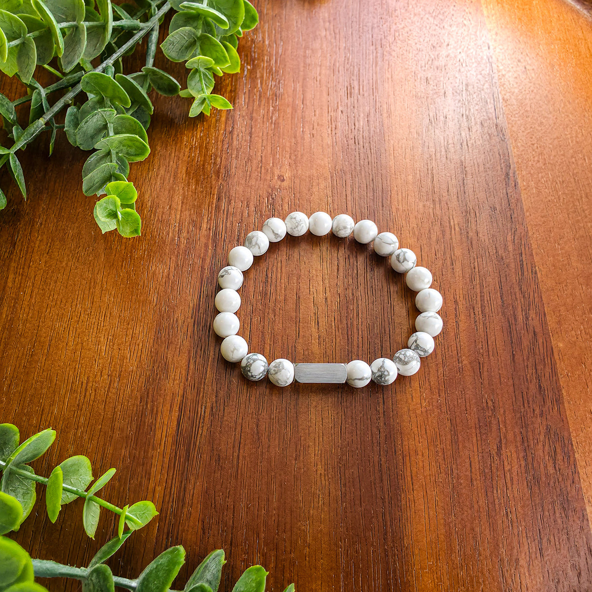 A beaded stretch bracelet made of white and gray marbled howlite beads with a silver rectangular bar for engraving, displayed on a warm brown wooden surface with green leaves in the corners.