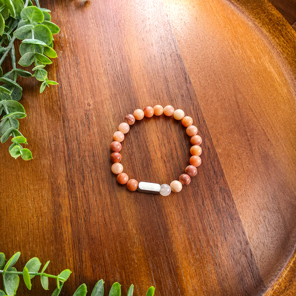 A beaded stretch bracelet with warm-toned round beads in shades of orange, terracotta, and cream, featuring silver rectangular bar for engraving, displayed on a wooden surface with green leaves in the corners.