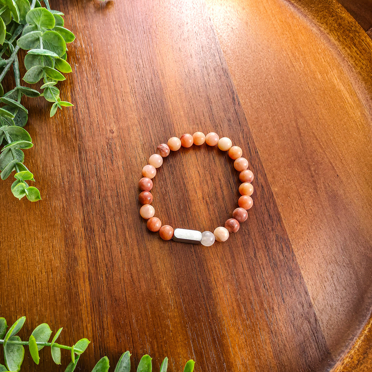 A beaded stretch bracelet with warm-toned round beads in shades of orange, terracotta, and cream, featuring silver rectangular bar for engraving, displayed on a wooden surface with green leaves in the corners.
