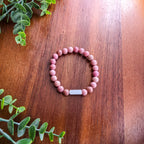 A beaded stretch bracelet made of pinkish-red round beads featuring silver rectangular bar for engraving, displayed on a wooden tabletop with green  leaves in the corners.
