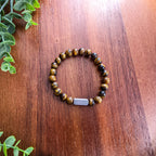 A beaded stretch bracelet made of round brown and black tiger's eye beads with a silver rectangular bar for engraving, displayed on a wooden tabletop with green leaves in the corner.