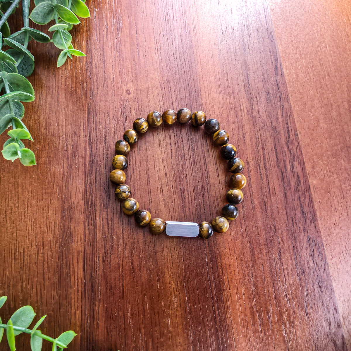 A beaded stretch bracelet made of round brown and black tiger's eye beads with a silver rectangular bar for engraving, displayed on a wooden tabletop with green leaves in the corner.