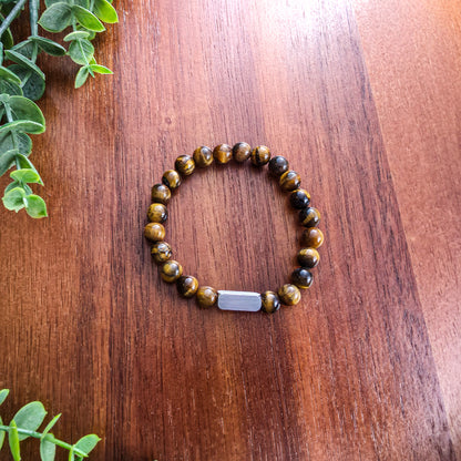 A beaded stretch bracelet made of round brown and black tiger's eye beads with a silver rectangular bar for engraving, displayed on a wooden tabletop with green leaves in the corner.