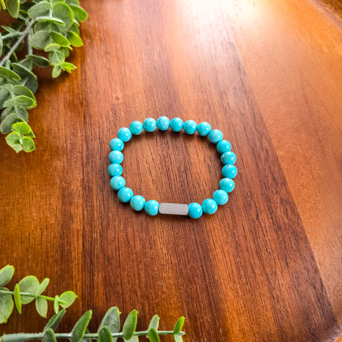 A turquoise beaded stretch bracelet with a silver rectangular bar for engraving, displayed on a polished wooden tabletop with green leaves in the corners.
